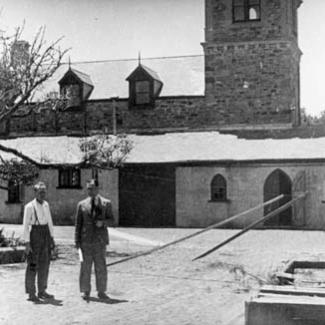A black and white photograph of the stone-masonry rear of St Francis House. There is a large, wilting tree and two people are standing in front of the tree.
