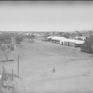 A black and white photograph of Mount Margaret Mission. There are several small buildings scattered among trees, with a large open clearing in front of the buildings. There is a person riding their bicycle in the clearing.
