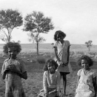 A black and white photograph of a group of children at Mount Margaret Mission. Some children are seated on the grass, and some are standing. They are smiling and looking at the photographer.