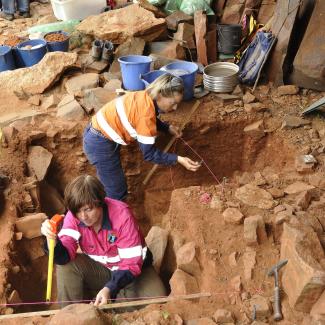 Two women measure the dimensions of a dig-site whilst a man sits beside them taking notes.